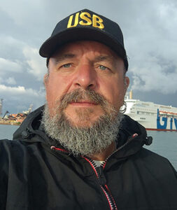 En man i grått stort skägg och med keps sår i en hamn. 