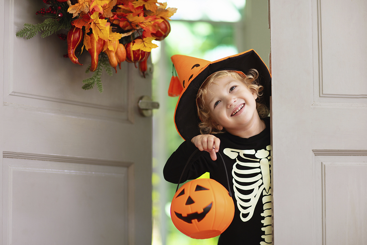 Kids trick or treat on Halloween night. Child at decorated house door with autumn leaf wreath and pumpkin lantern. Little boy in witch and skeleton costume and hat with candy bucket. Fall decoration.
